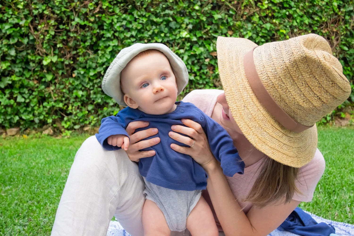 Madre sosteniendo a su bebé en un entorno al aire libre, con césped verde y plantas de fondo. El bebé lleva un sombrero claro y una camiseta azul, mientras que la madre usa un sombrero de paja. Ambos muestran una expresión de alegría y conexión.