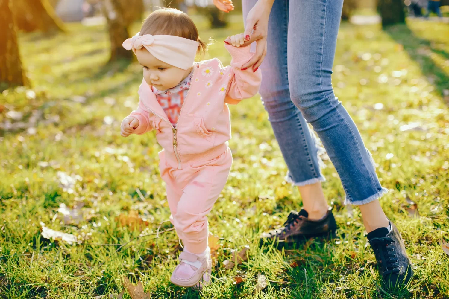 Niña pequeña vestida con un conjunto rosa y una diadema, caminando en un parque mientras sostiene la mano de un adulto. El entorno muestra hierba verde y árboles al fondo, iluminado por la luz del sol.