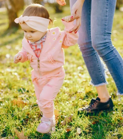Niña pequeña vestida con un conjunto rosa y una diadema, caminando en un parque mientras sostiene la mano de un adulto. El entorno muestra hierba verde y árboles al fondo, iluminado por la luz del sol.