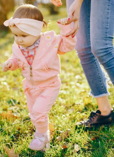 Niña pequeña vestida con un conjunto rosa y una diadema, caminando en un parque mientras sostiene la mano de un adulto. El entorno muestra hierba verde y árboles al fondo, iluminado por la luz del sol.
