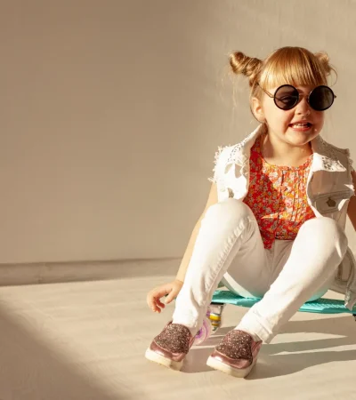 Niña sonriente sentada sobre una patineta turquesa, vestida con camiseta colorida y chaleco blanco, usando gafas de sol y zapatillas brillantes, en un ambiente luminoso y moderno.