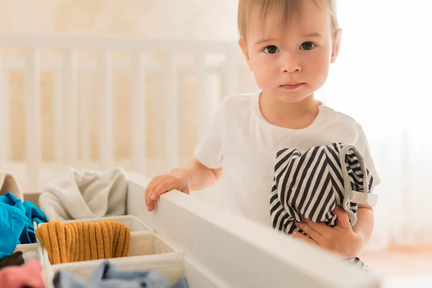 Niño pequeño sosteniendo una prenda de ropa a rayas, frente a un cajón lleno de ropa de colores variados, en un ambiente luminoso y acogedor.