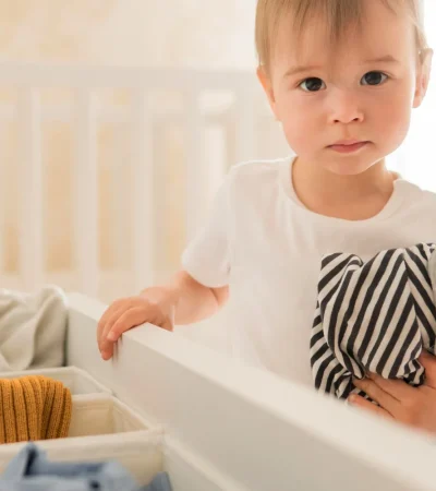 Niño pequeño sosteniendo una prenda de ropa a rayas, frente a un cajón lleno de ropa de colores variados, en un ambiente luminoso y acogedor.