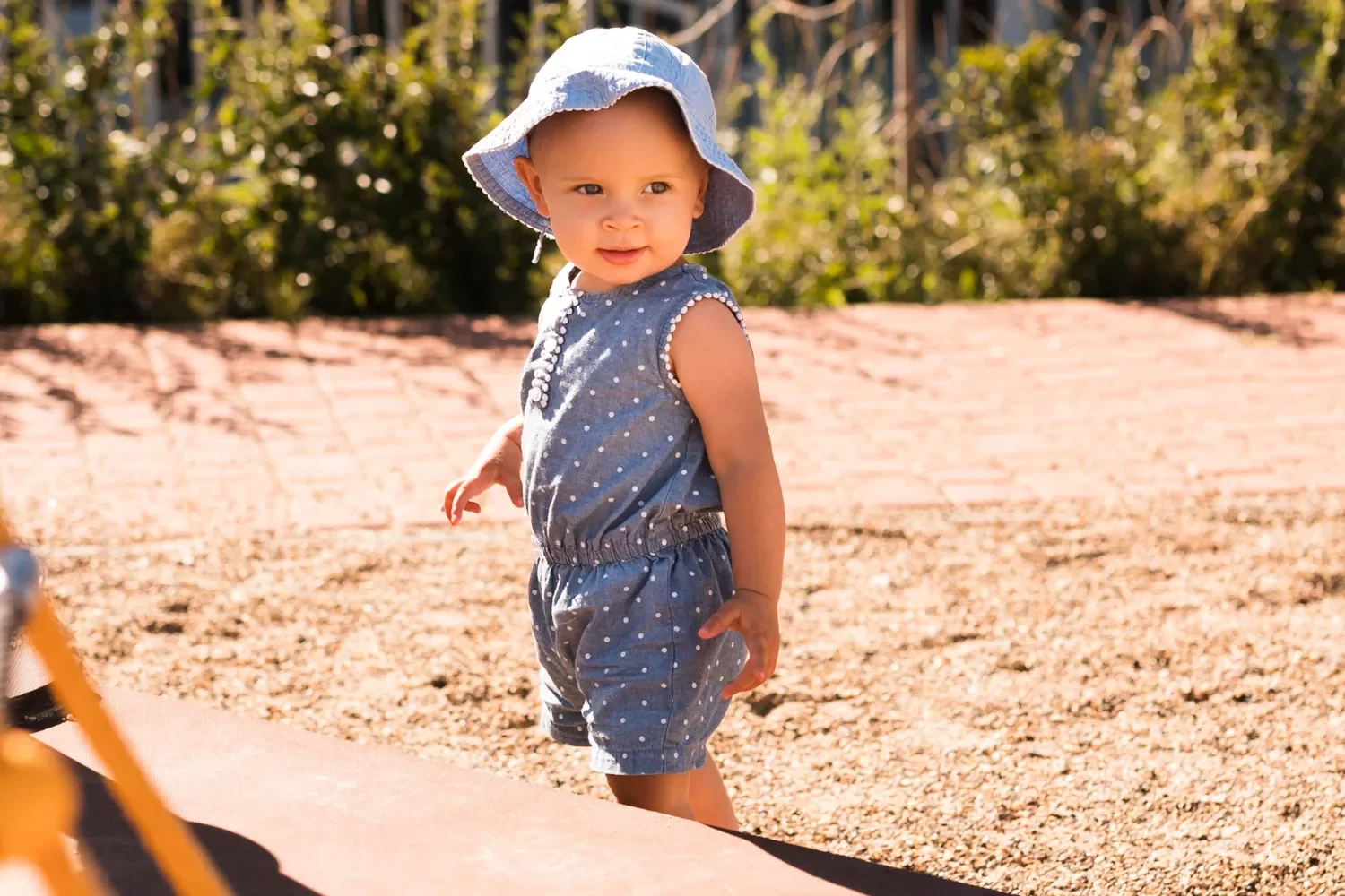 Niña pequeña sonriente con sombrero azul claro, vestida con un overol de mezclilla con lunares, jugando en un parque soleado. Fondo de arena y vegetación.