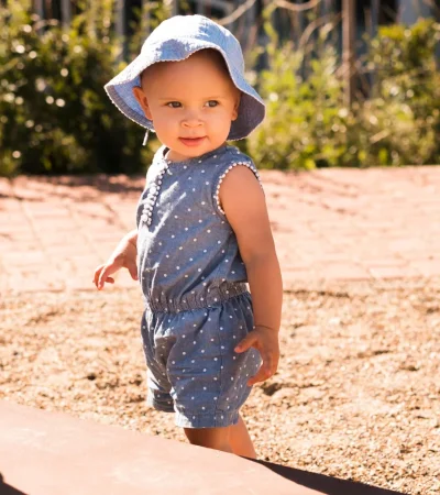 Niña pequeña sonriente con sombrero azul claro, vestida con un overol de mezclilla con lunares, jugando en un parque soleado. Fondo de arena y vegetación.