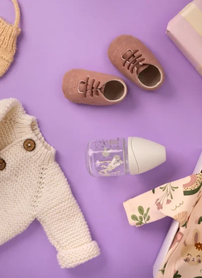 Conjunto de ropa para bebé que incluye un suéter blanco de punto, un body con estampado floral, un chupete, zapatos de cuero rosa, mitones de lana y un regalo envuelto en papel rosa con un lazo. Fondo morado que resalta los artículos.