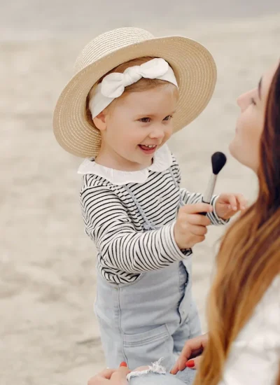 Niña pequeña con sombrero de paja juega a maquillarse con su madre en la playa, mostrando un momento de alegría y conexión familiar.