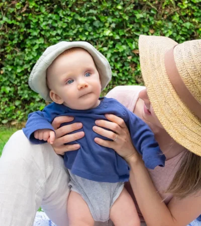 Madre sosteniendo a su bebé en un entorno al aire libre, con césped verde y plantas de fondo. El bebé lleva un sombrero claro y una camiseta azul, mientras que la madre usa un sombrero de paja. Ambos muestran una expresión de alegría y conexión.
